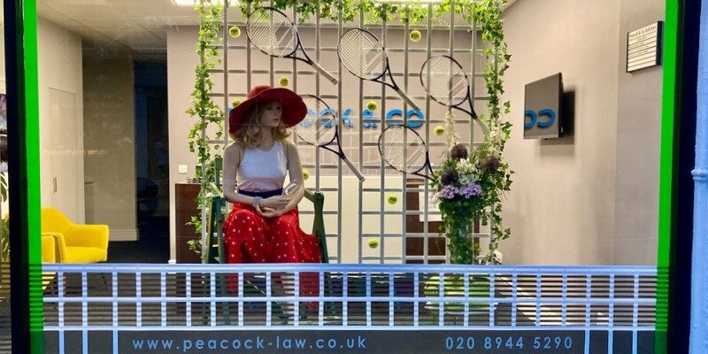 Peacock & Co Wimbledon Championships window display of mannequin in red hat and skirt, white top and tennis racquets on a trellis.