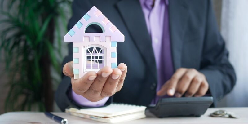 A man holding a small model house in his hand, while using a calculator and notebook in his other, signifying financial impact of the Stamp Duty Land Tax cuts announced in the Chancellor's mini bugdet.