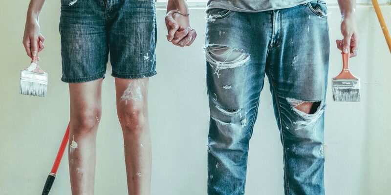 Male in ripped jeans and female in cut-off jean shorts with paintbrushes signifying first-time buyers of a property purchase.